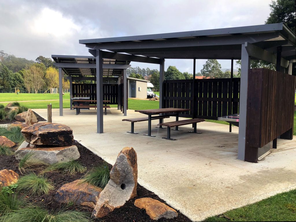 Two covered picnic areas with wooden tables and benches on a concrete slab. Surrounding the area are large rocks and grassy landscaping. A large open field and trees are visible in the background under a cloudy sky.