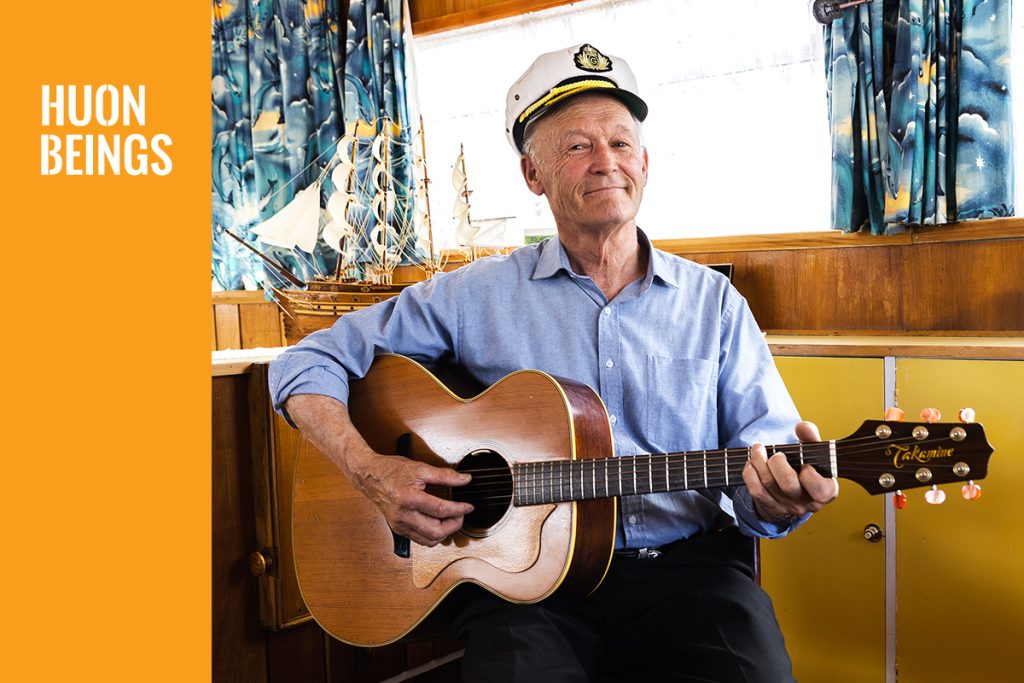 Elderly man wearing a captains hat smiles as he plays an acoustic guitar. He sits indoors, with a model ship and curtains in the background. The text HUON BEINGS is displayed on the left side in white on an orange background.