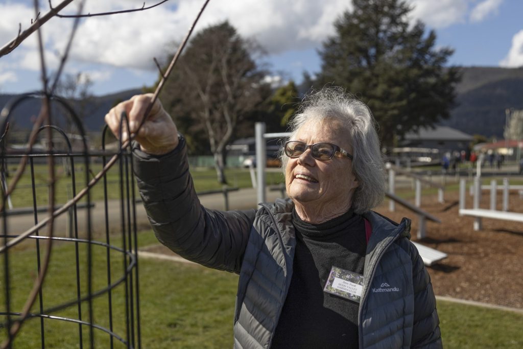 An elderly woman with gray hair and sunglasses carefully tends to a plant in a park. Shes wearing a dark jacket and has a name tag on her chest. In the background, there are trees and a partly cloudy sky.