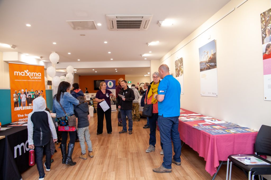 A group of people are gathered in a room with wooden floors and bright lighting, engaging in discussions at a job fair. Tables with brochures and banners from various companies are set up along the walls.