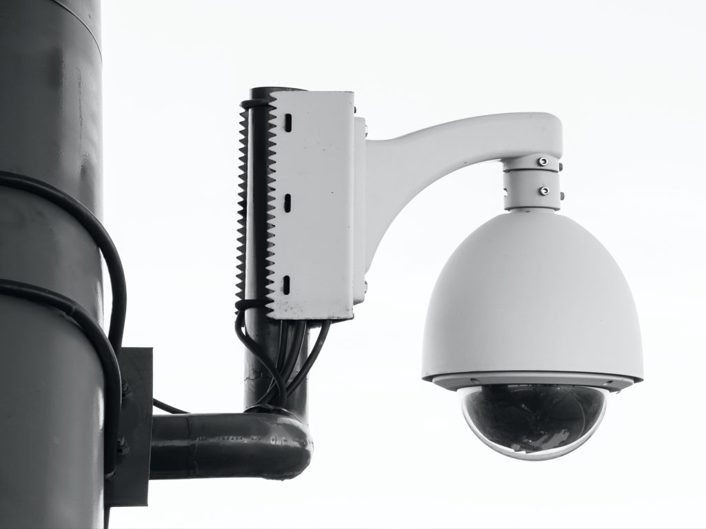 A white security camera with a dome shape is mounted on a pole. The background is a clear, overcast sky. The camera is surrounded by metal brackets and wiring.