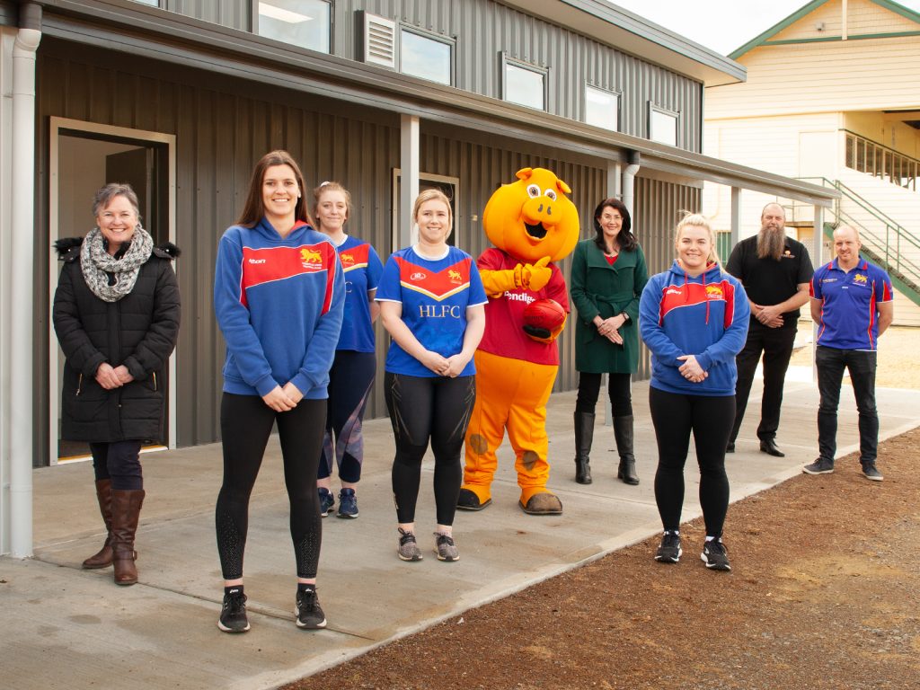 A group of people, including four women in sports jerseys and a person in a mascot costume, stand outside a building. They are arranged in a line, smiling at the camera, with a metal-clad building in the background.
