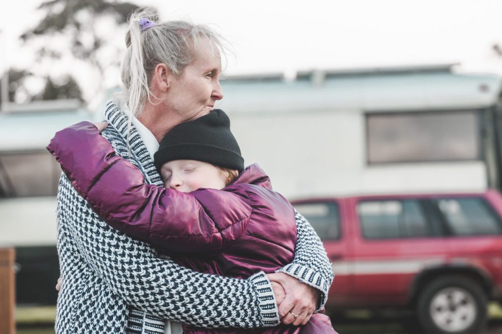 A woman in a patterned coat gently hugs a child in a purple jacket and black beanie. The child has their eyes closed. A red vehicle and a caravan are visible in the background.
