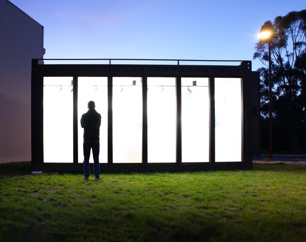 Silhouette of a person standing in front of a brightly lit building with large glass panels at dusk. The sky is a deep blue, and a streetlight illuminates a patch of grass. Trees are visible in the background.