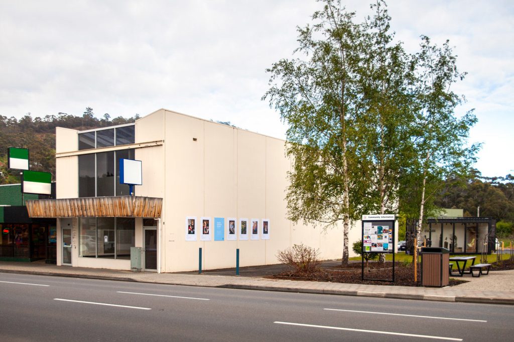 A modern, white building with large windows is situated next to a small green shop. Posters are displayed on the buildings side. A tree and a map stand are on the sidewalk, along with a bench and trash can. A road runs in front.