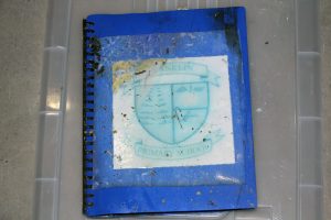 A worn blue spiral notebook with a logo for Franklin Primary School on the cover, resting on a transparent plastic surface. The cover has dirt marks and signs of wear.