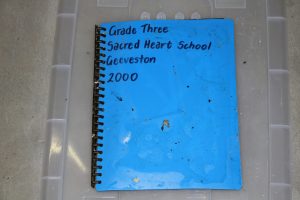 A blue spiral-bound notebook is labeled with Grade Three, Sacred Heart School, Geeveston, 2000 handwritten in black ink. The notebook is placed on a clear plastic surface.