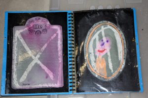 A worn spiral-bound book is open to two pages. The left page has a Me title with smiley faces and is scribbled over with a large X. The right page shows a childs drawing of a smiley face in a circular frame.