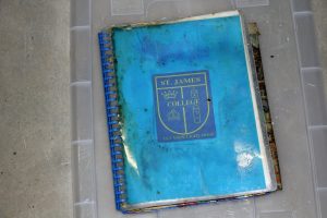 A worn blue spiral-bound notebook on a transparent plastic surface. The notebooks cover features a St. James College emblem with a crown, book, and pen design, and the motto Let Your Light Shine below the emblem.