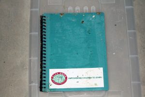 A worn, blue spiral notebook with a Yelin Hui Primary School logo and the slogan Empowering Children to Learn is placed on a clear plastic surface. The cover shows signs of wear and scattered dirt.