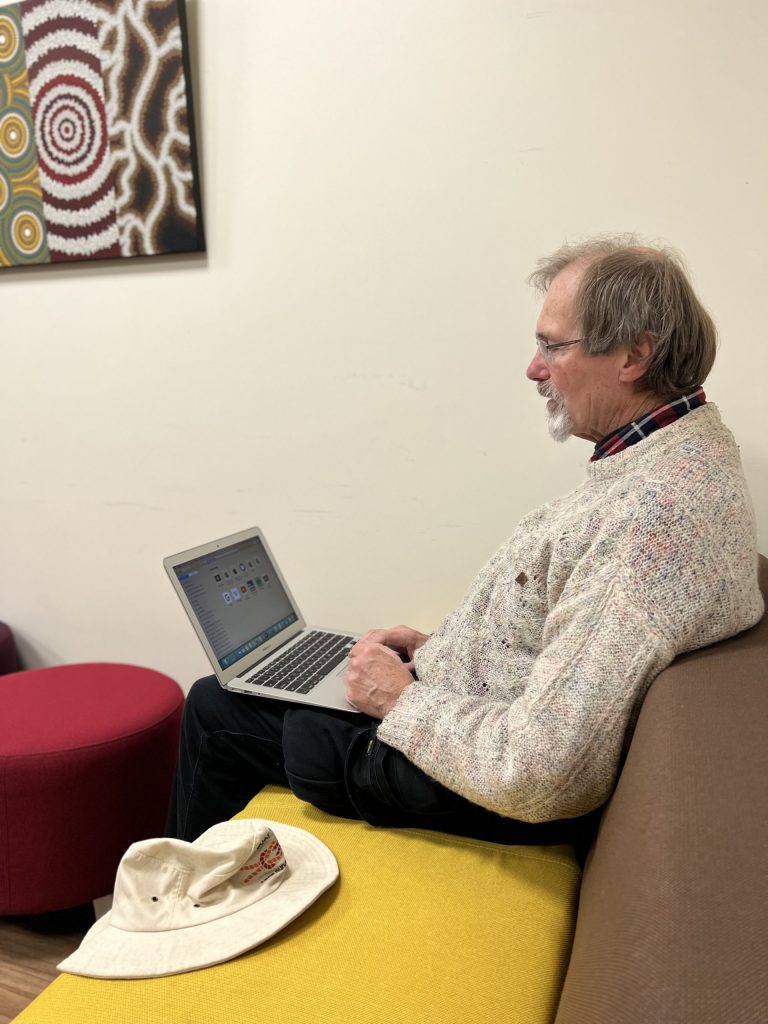 A man with glasses and a sweater is sitting on a couch using a laptop. A beige hat is beside him. Theres a colorful abstract painting on the wall and a red cushioned stool nearby.