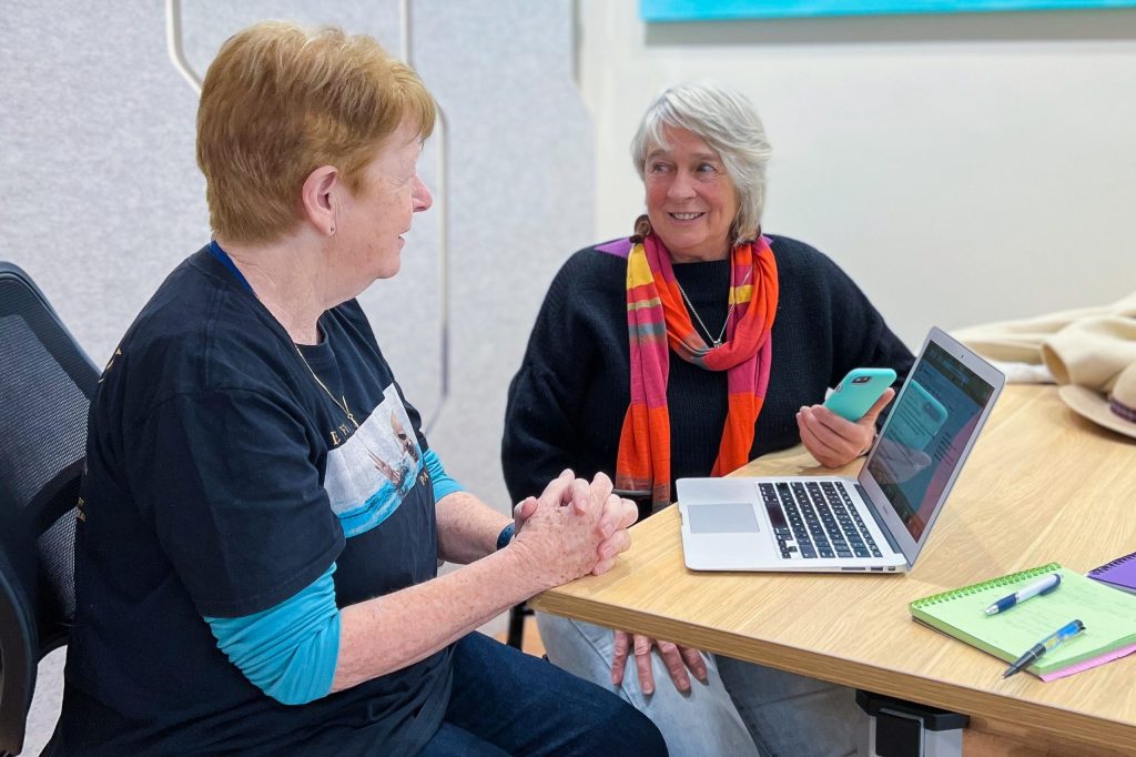 Two women sit at a table with a laptop, notebook, and pen. One woman is holding a smartphone and smiling. They appear engaged in conversation. Both are casually dressed, and the setting is a bright, modern room.