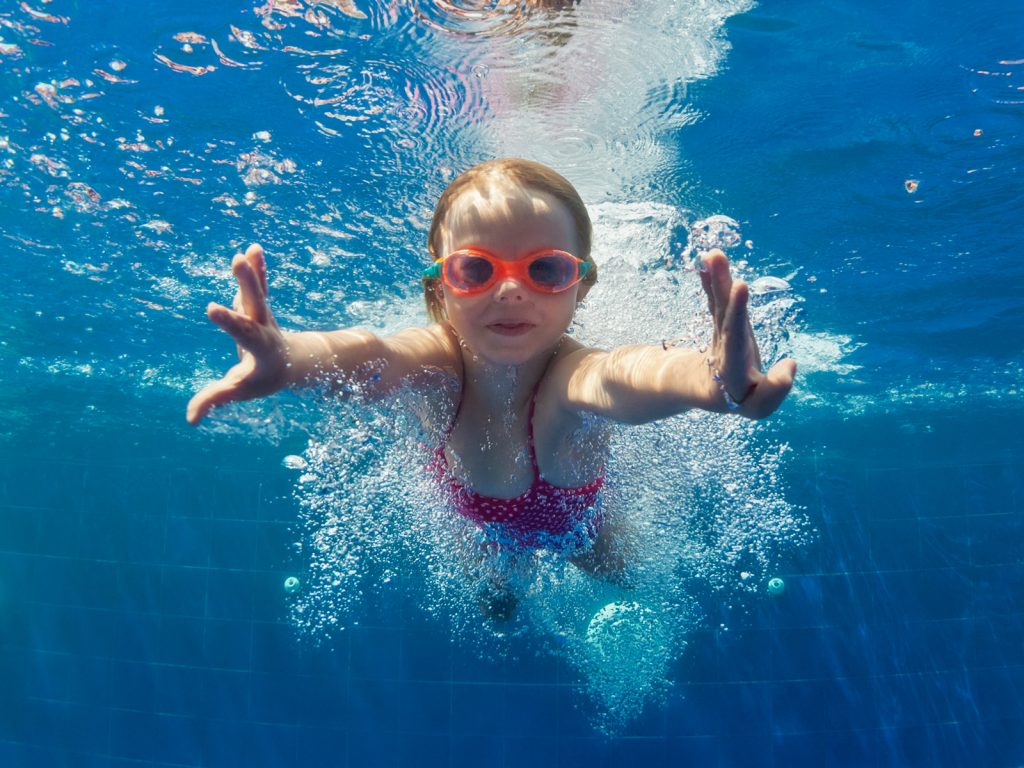 A child swims underwater in a pool, reaching toward the camera with both hands. She wears red goggles and a pink swimsuit. Bubbles trail around her, and sunlight shimmers through the blue water.