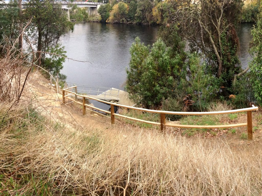 A narrow, unpaved path with a wooden railing leads down a grassy slope to a calm river. The scene is surrounded by trees and bushes, with a bridge visible in the background over the water.