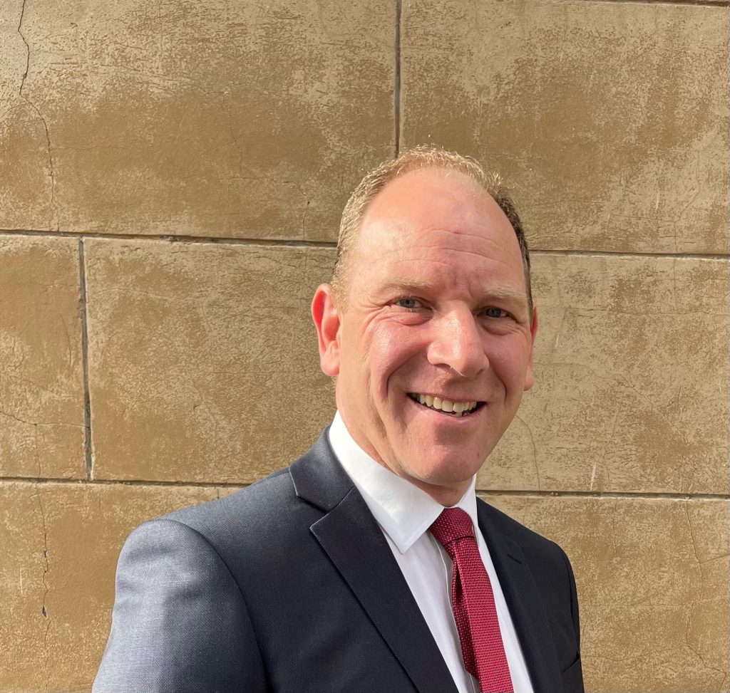 A man in a dark suit and red tie smiles while standing in front of a textured, stone wall. The lighting is bright, casting slight shadows.
