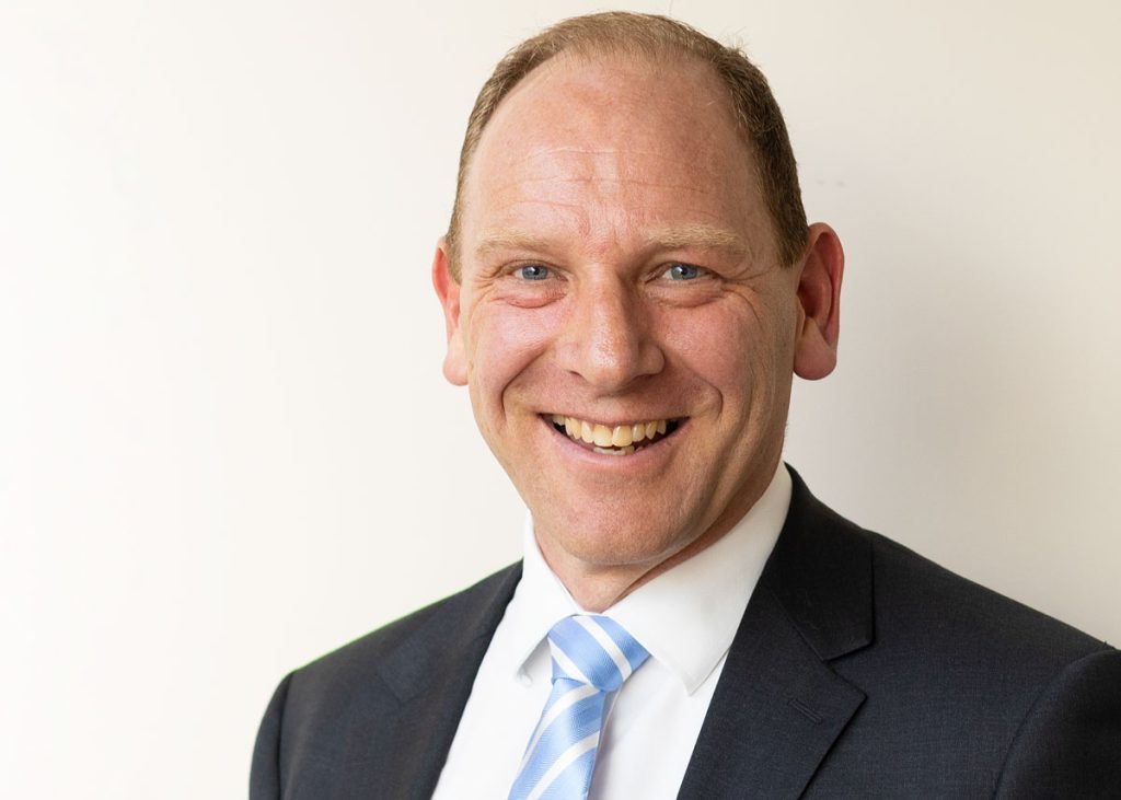 A smiling man in a dark suit and light blue striped tie is standing against a plain white background. He has short, light brown hair and blue eyes.