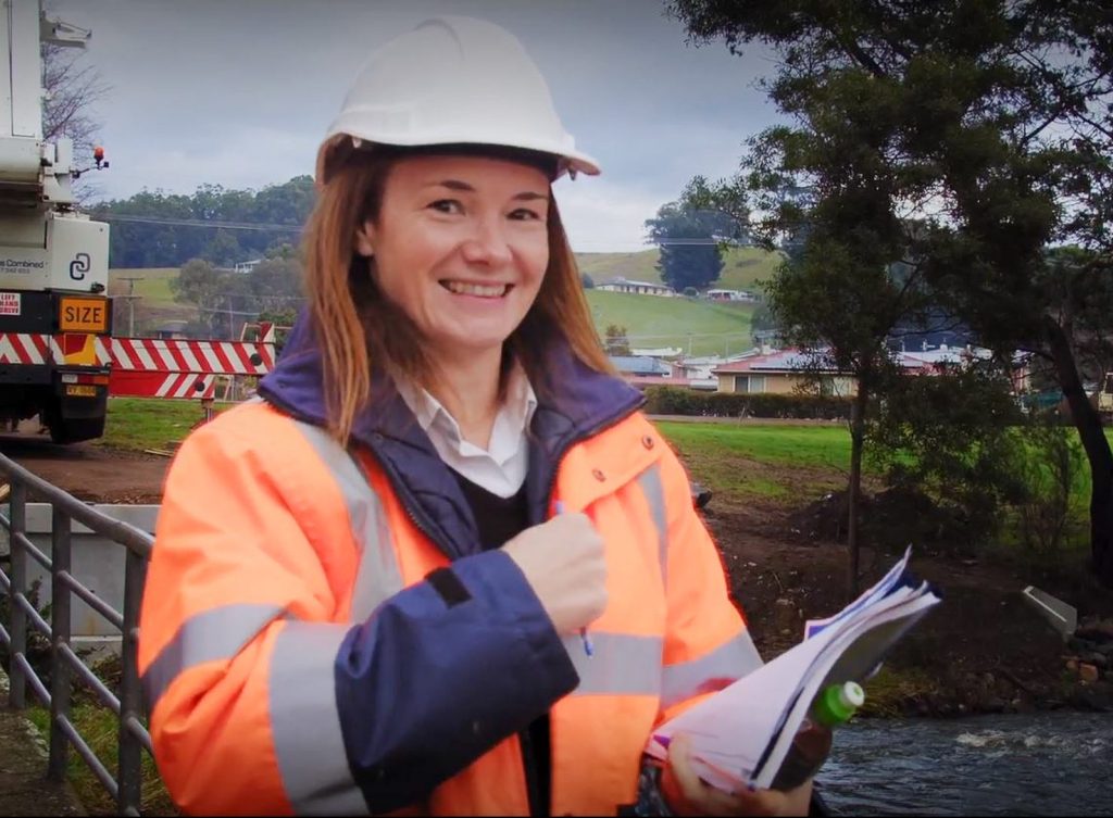 A person in an orange reflective jacket and white hard hat stands outdoors, smiling and giving a thumbs up. They are holding papers in their other hand. Trees and a construction vehicle are in the background.