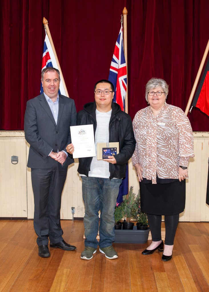 Three people stand indoors on a wooden floor in front of red curtains. The person in the middle holds a certificate and a small flag plaque. Two flags are displayed behind them. The group is smiling at the camera.