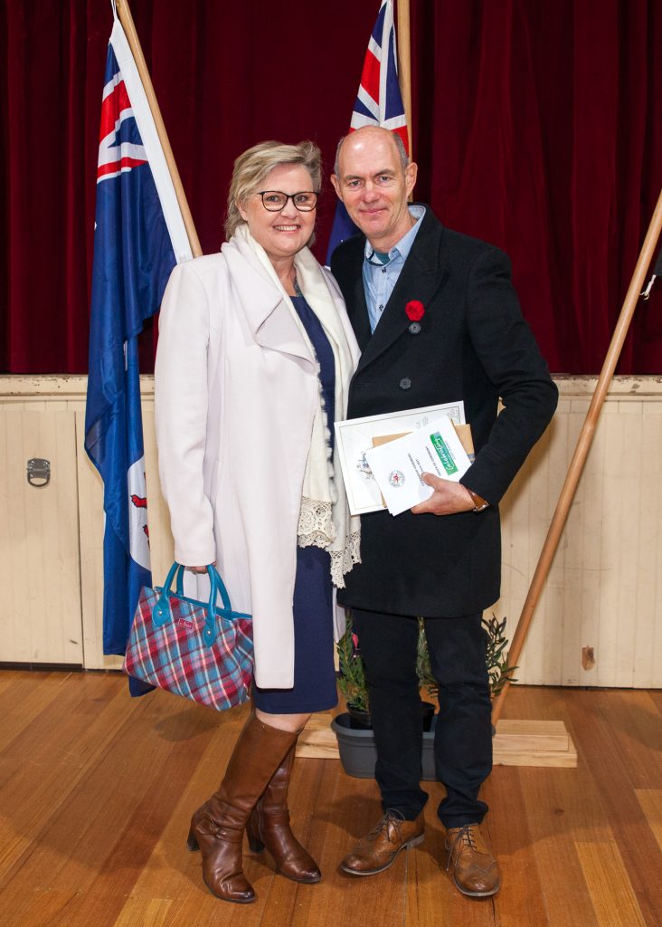 A woman in a white coat and boots stands beside a man in a dark coat holding papers, in front of red curtains and flags. They are on a wooden floor, and the man has a poppy pinned to his coat.