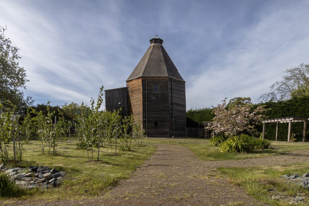 A tall, wooden, hexagonal building stands at the end of a dirt path, surrounded by a grassy area with small trees and flowering bushes. The sky is partly cloudy, and there are hedges in the background.