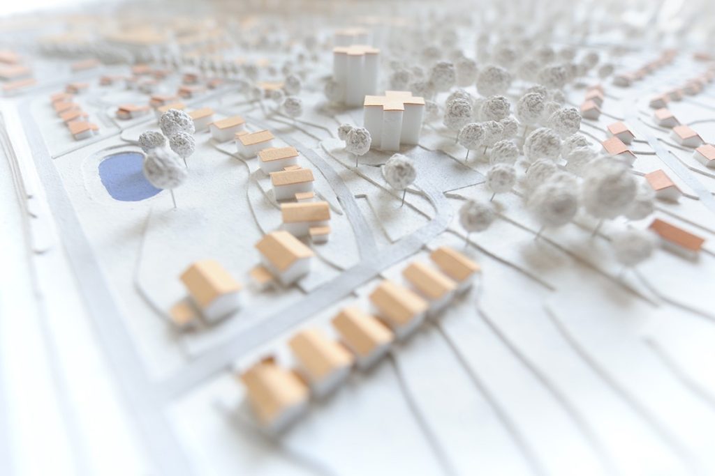 A miniature diorama showing a suburban neighborhood with small houses featuring brown roofs, a pond, trees, and a few larger buildings with cross-shaped roofs amidst other structures, all set on a textured landscape.