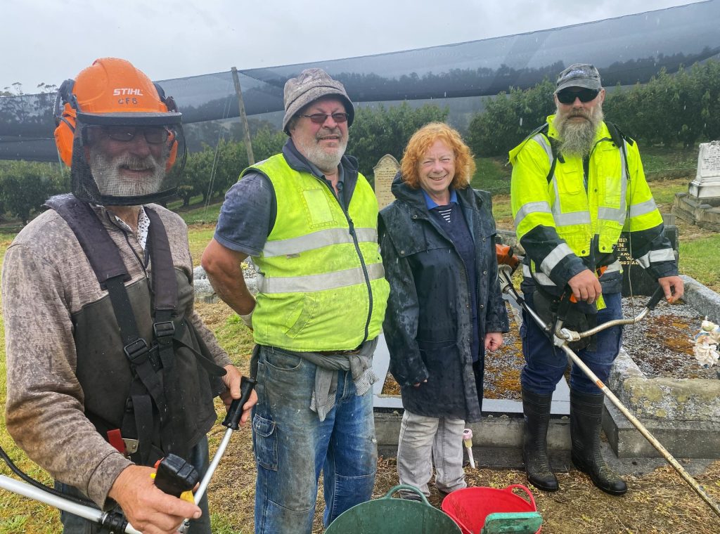 Four people stand outdoors in a grassy area. Two are holding string trimmers, and three wear reflective vests. They are smiling, and the background includes a fence and trees. Its a cloudy day.