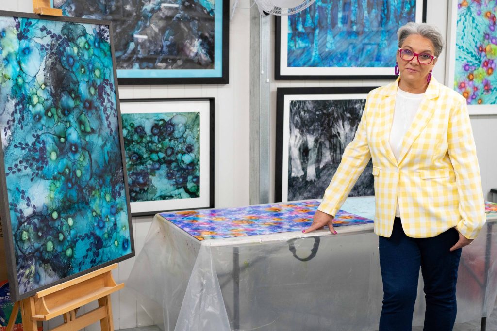 A person with short gray hair and glasses, wearing a yellow checkered blazer, stands in an art studio. They are posing with colorful abstract paintings displayed on the walls and a table.