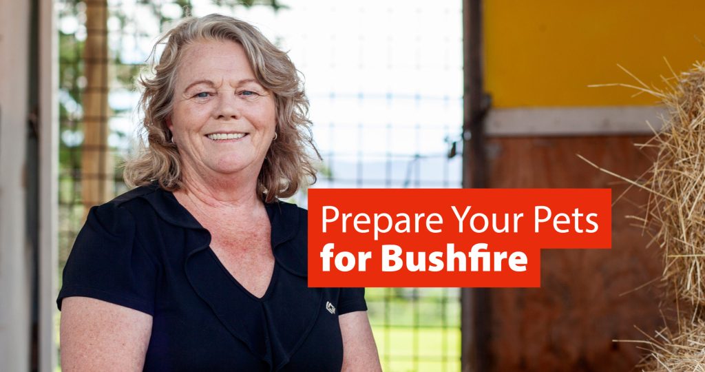 A smiling woman with wavy, shoulder-length hair stands indoors near hay bales. She is wearing a black shirt. A red sign next to her reads, Prepare Your Pets for Bushfire. The background includes a metal mesh and yellow wall.