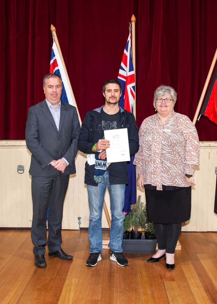 Three people stand indoors on a wooden floor with maroon curtains and flags behind them. The person in the middle holds a certificate. The two others, a man in a suit and a woman in a patterned blouse, stand on either side, smiling.