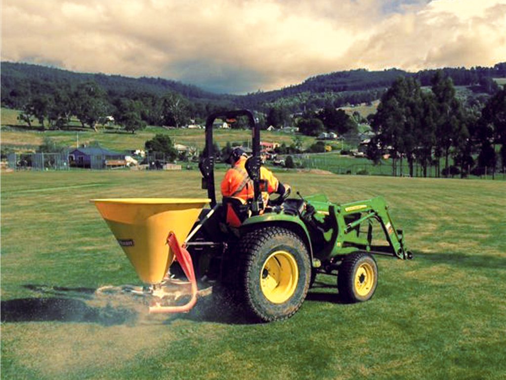 A person operating a green tractor with a yellow fertilizer spreader attachment on a large grassy field. They are wearing an orange safety vest and helmet. Trees and hills are visible in the background under a cloudy sky.