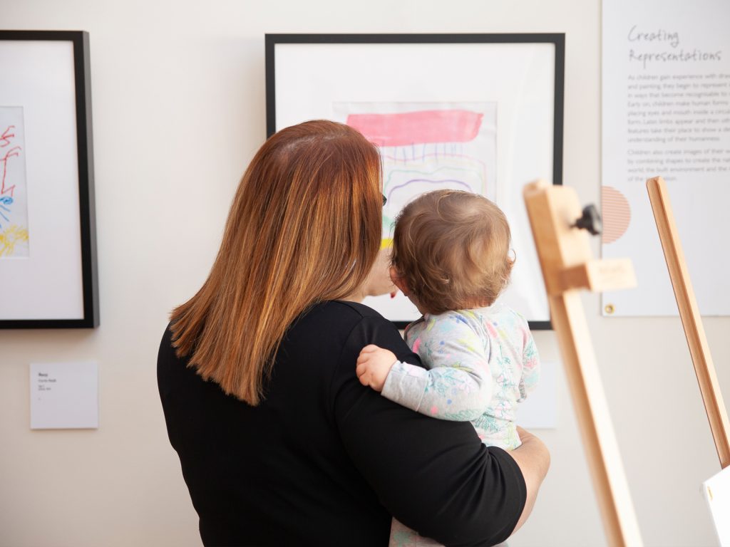 A woman with auburn hair is holding a young child in her arms. They are viewing colorful abstract artwork framed on a gallery wall. The scene suggests a nurturing interaction and an interest in art appreciation.