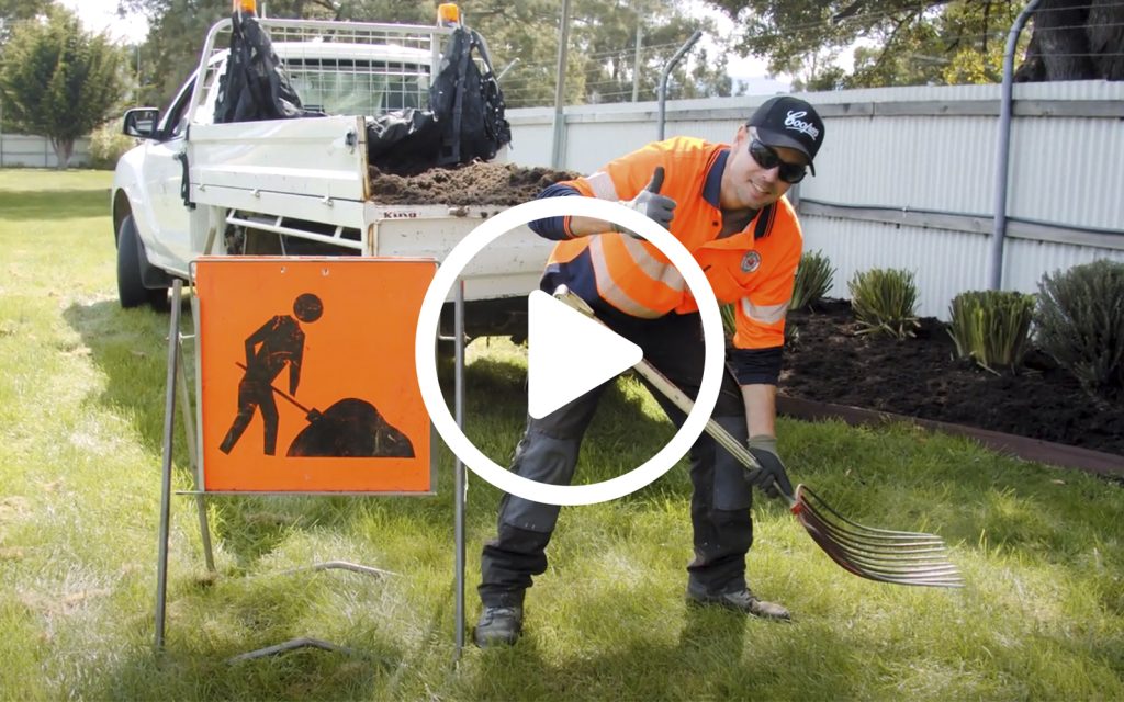 A person wearing an orange safety vest and cap gives a thumbs-up while holding a pitchfork. They are next to a work sign and a truck loaded with dirt. The scene is set in a grassy area with trees and shrubs in the background.