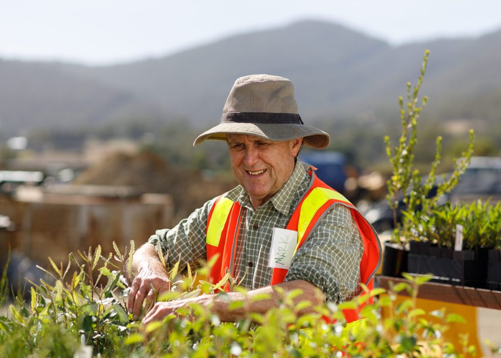 An older adult wearing a wide-brimmed hat, plaid shirt, and orange safety vest tends to plants outdoors. Mountains are visible in the background. A name tag on the vest reads Mark.