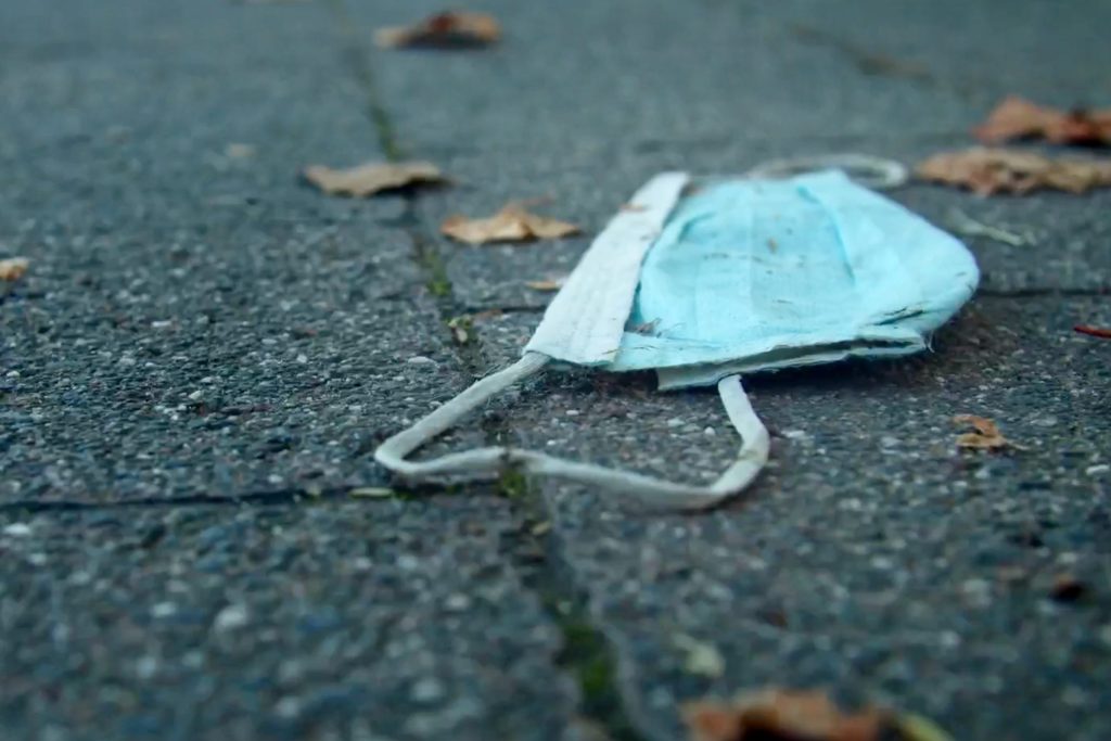 Discarded blue surgical mask lying on a pavement with scattered dried leaves. The ground is made of grey stone, and the scene suggests neglect or pollution.