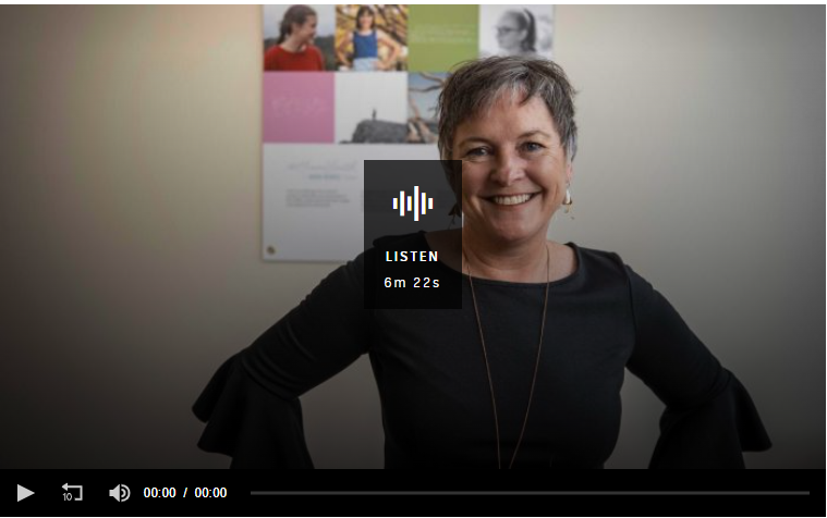 A person with short hair smiles while standing with hands on hips in an office setting. They are wearing a black top. Behind them is a blurred bulletin board with photos and papers. Audio player overlay with play duration shows LISTEN 6m 22s.