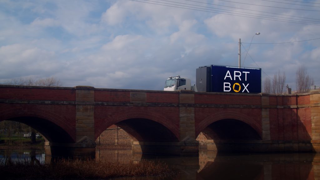 A truck with a blue container labeled ART BOX drives over a stone bridge with three arches. The bridge is reflected in the water below. The sky is cloudy, and power lines are visible above.