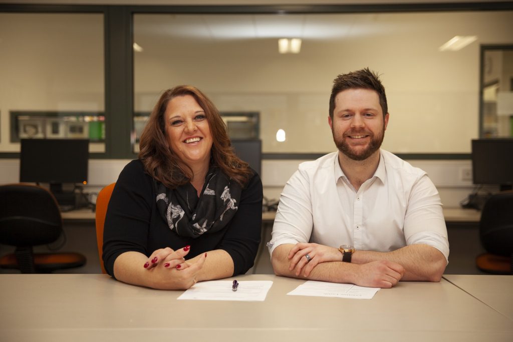 Two people sitting at a table in an office setting, smiling at the camera. There are documents on the table and computers in the background. The woman is on the left, wearing a black scarf, and the man is on the right, in a white shirt.