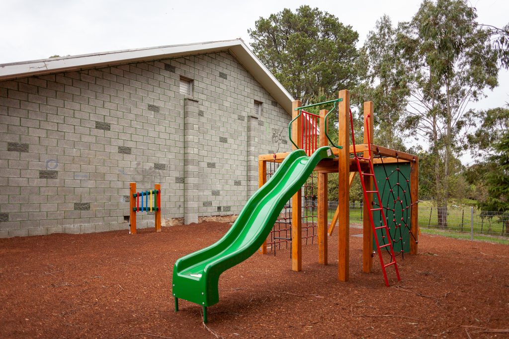 A playground with a green slide, red ladder, and climbing structure is set on a bark-covered ground next to a brick building. Trees and a fence are in the background.