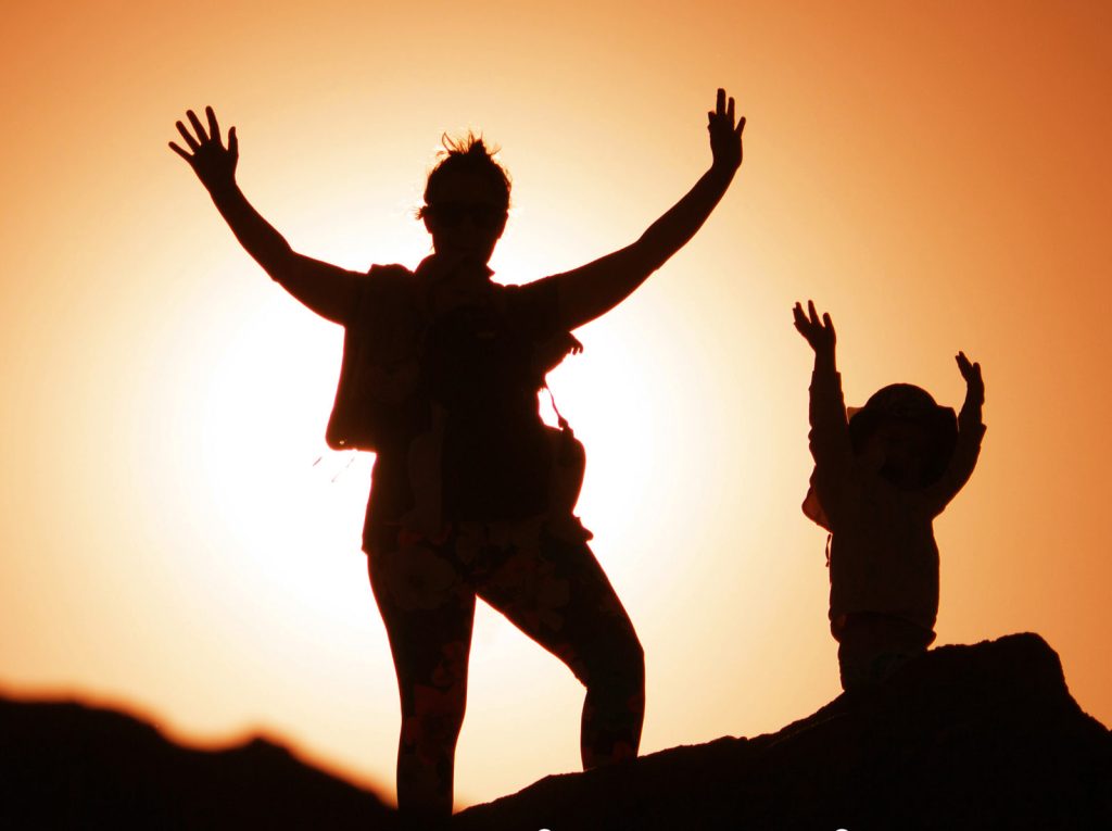 Silhouette of an adult and child standing on rocks against a setting sun. Both have their arms raised in celebration or greeting, with an orange sky creating a warm backdrop.