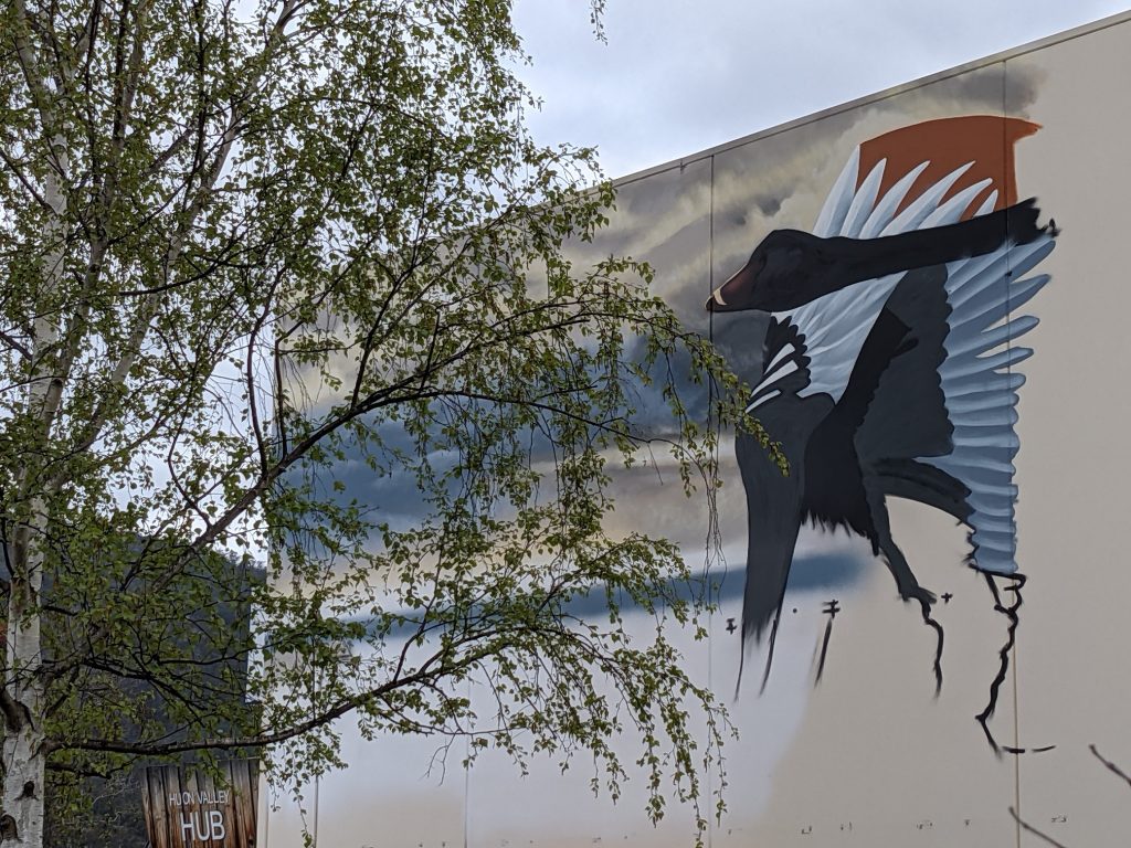 A mural of a bird with outstretched wings is painted on a building. The sky behind is cloudy. A tree with green leaves partially obscures the view on the left side. A small sign reading HUB is visible in the lower left corner.