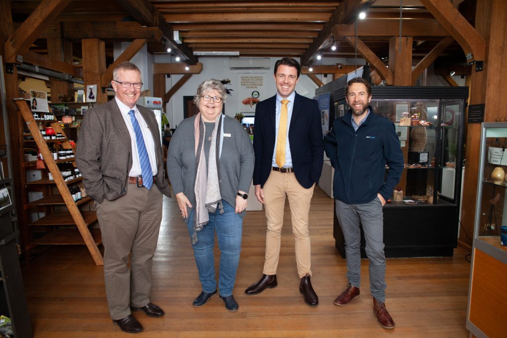 Four people stand smiling in a wooden interior space with shelves and displays. Two people are wearing suits, one is wearing a cardigan, and one is wearing a jacket and jeans. They are positioned in a staggered row.