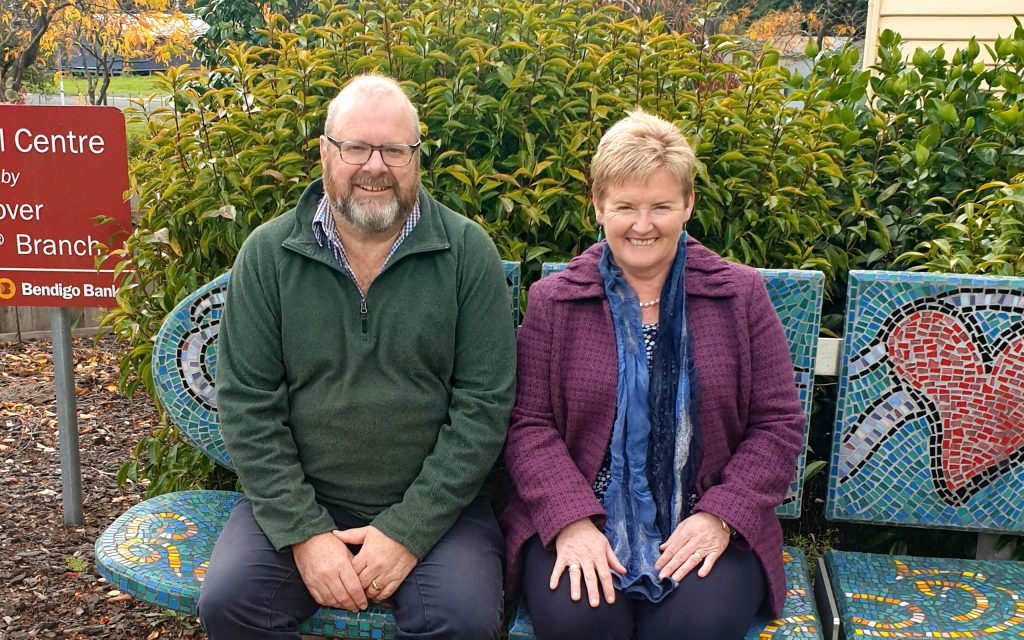 A man and woman are sitting on a colorful mosaic bench outdoors. The man wears a green jacket and the woman wears a purple coat and blue scarf. Theyre smiling, with green bushes and a red sign in the background.
