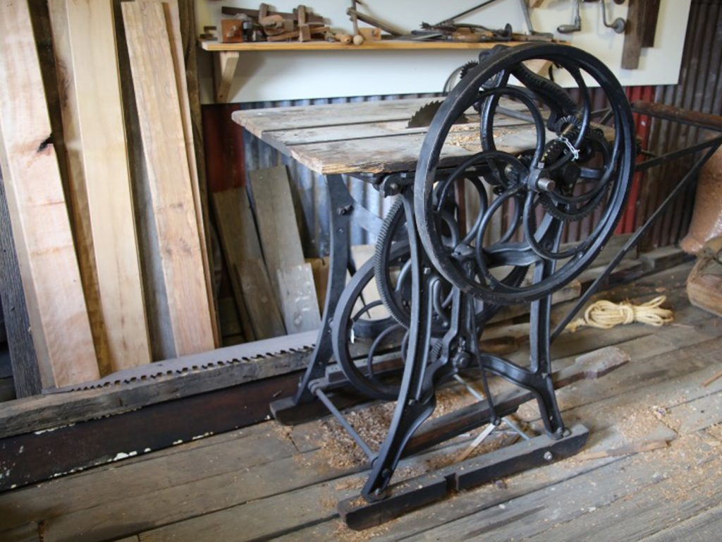 An old-fashioned sawmill machine with large metal wheels stands in a workshop. Wood planks and ropes are nearby. The floor is wooden, and various tools hang on the wall in the background.