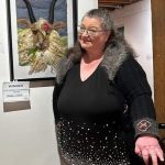 A woman smiling and gesturing with her hand stands next to award-winning artwork of a ram with textured fleece. The artwork is displayed in a frame on a gallery wall. A sign indicates it as the winner of an outstanding achievement award.