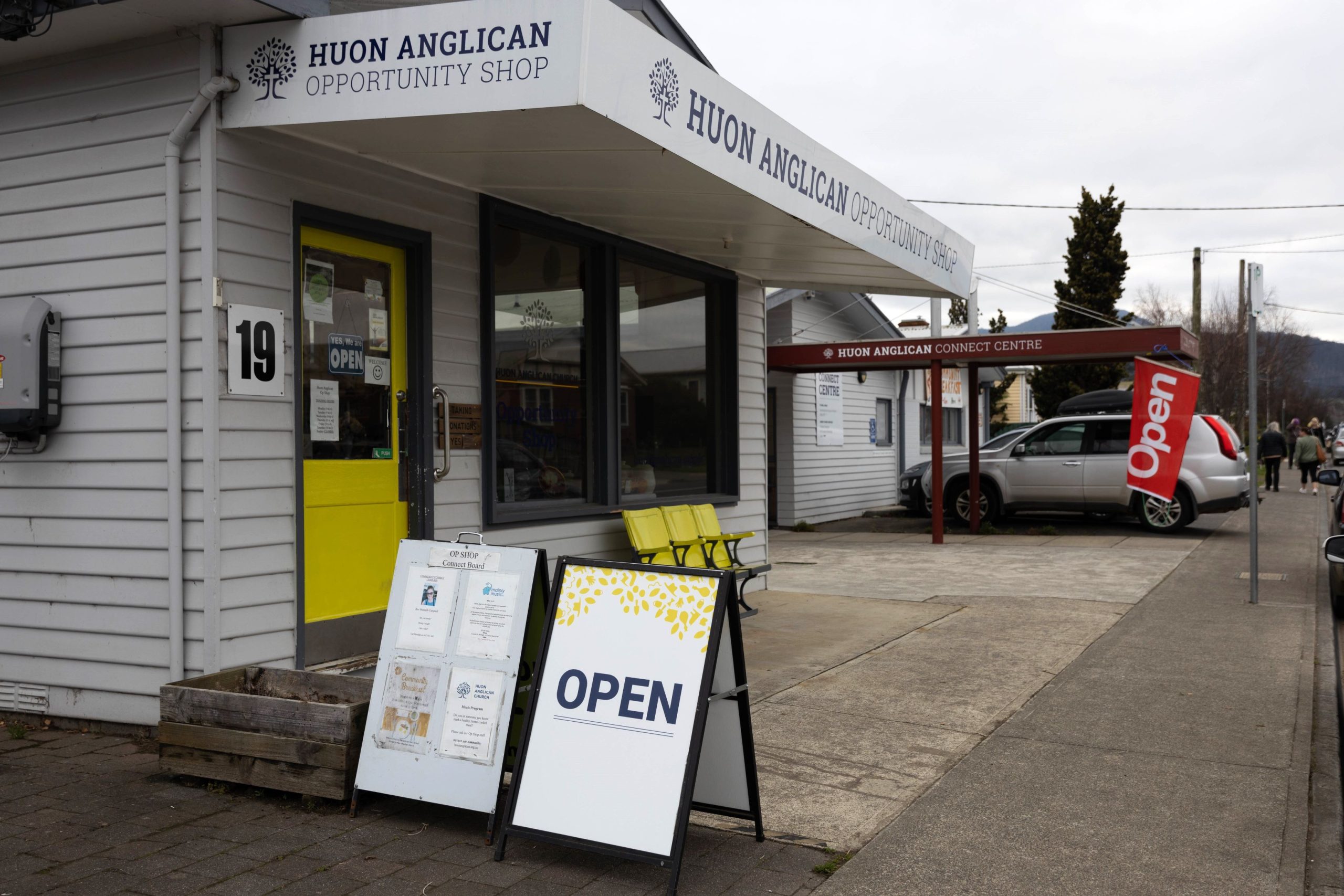 Exterior of Huon Anglican Opportunity Shop with a sign reading OPEN on the sidewalk. A yellow bench is by the window under an awning. Nearby is the Huon Anglican Community Centre, and a car is parked in the distance.