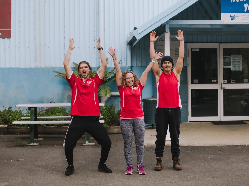 Three people in red shirts stand in front of a building, smiling with arms raised. Two men are on either side of a woman. They are outdoors, on a paved area, with a blue wall and glass door in the background. A bench with plants is on the left.