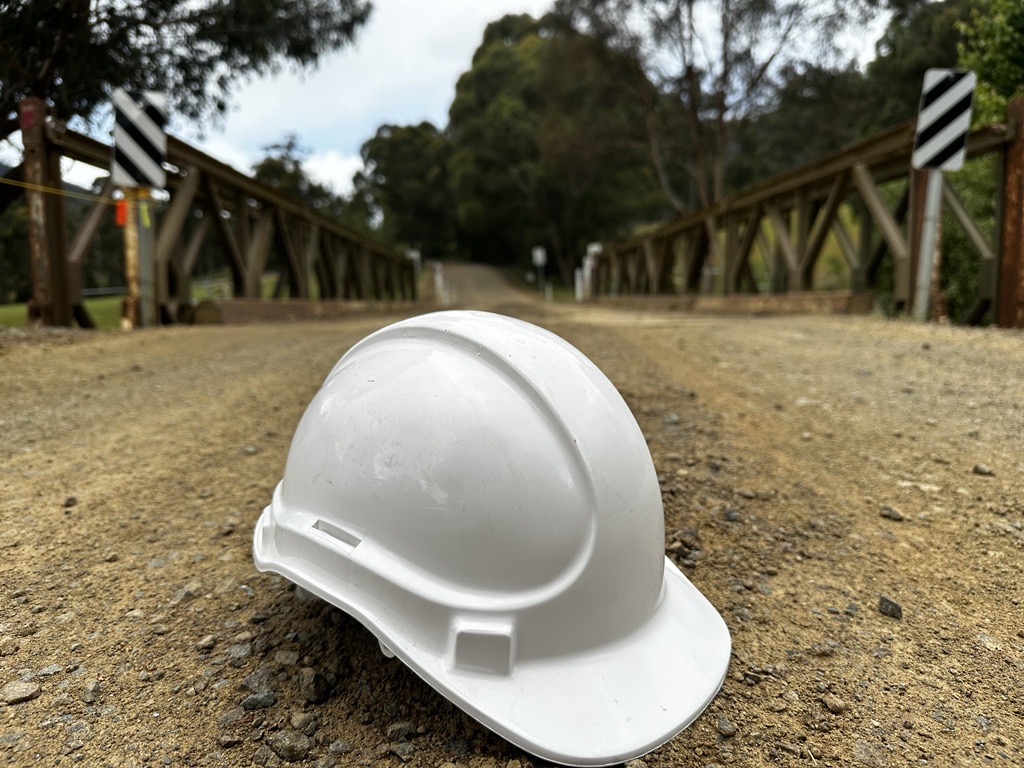 A white hard hat lies on a gravel road leading to a wooden bridge. The bridge has black and white striped signs on either side. Trees surround the area, and the sky is cloudy, creating a secluded atmosphere.