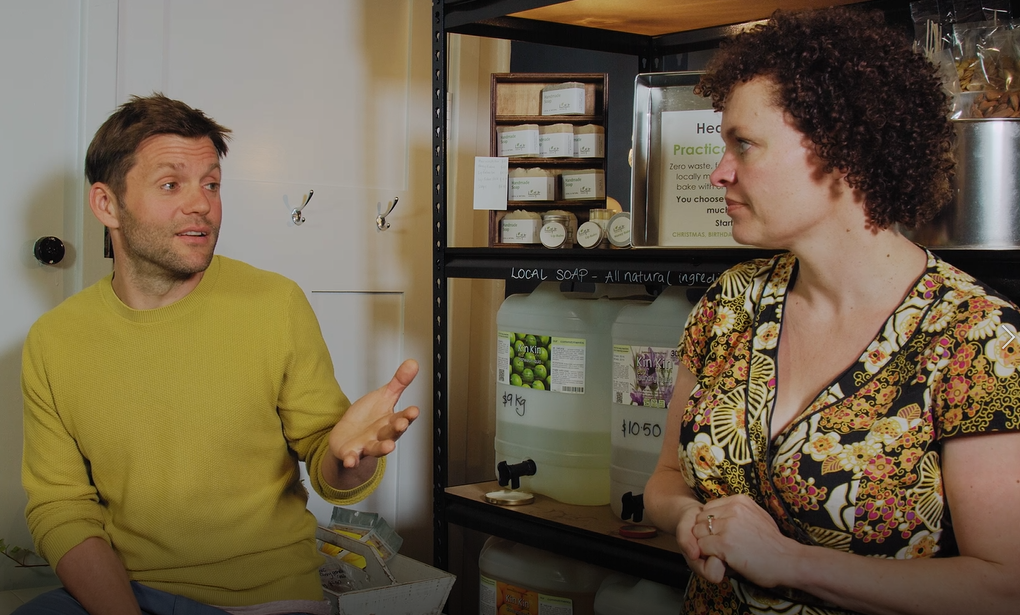 A man in a yellow sweater and a woman in a floral dress engage in conversation in front of a shelf stocked with soap and personal care products. The background features dispensers and product labels.
