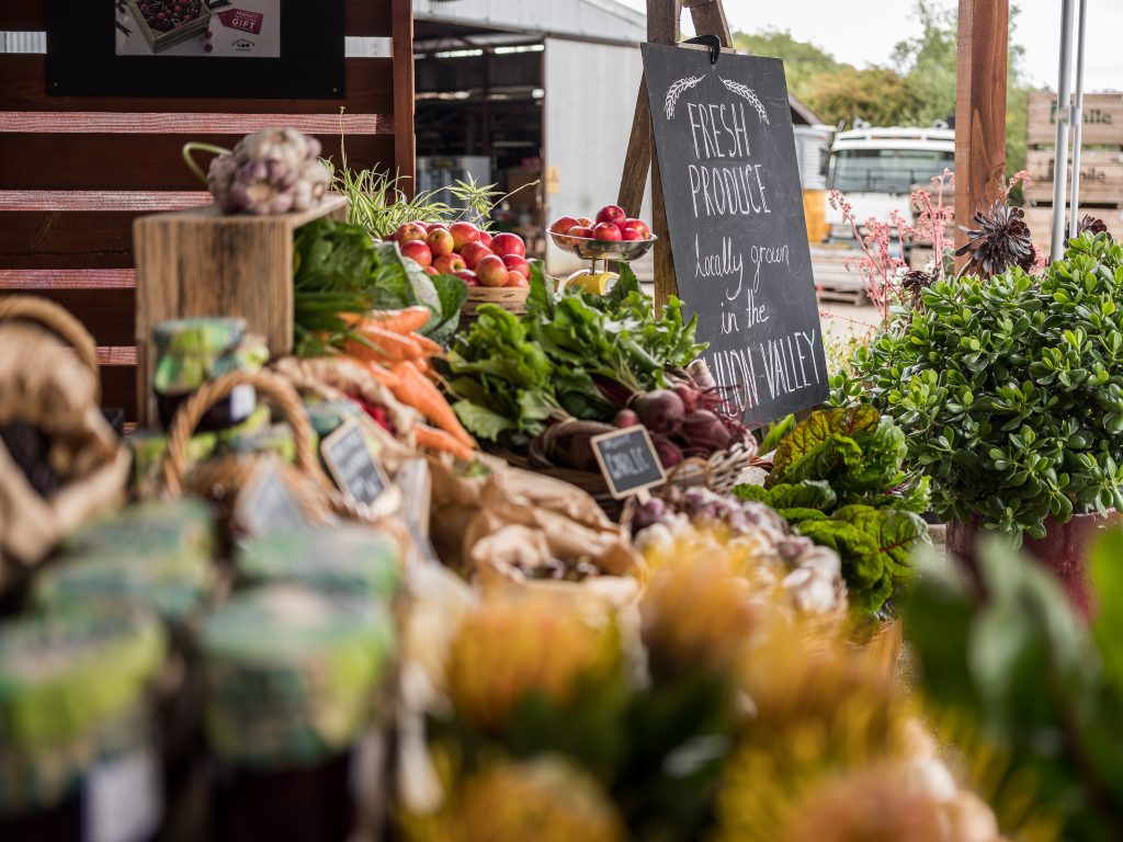 A display of fresh produce at a market stall, featuring carrots, apples, beets, and leafy greens. A chalkboard sign reads Fresh produce locally grown in the Yarra Valley. Jars of preserves and flowers are visible in the foreground.