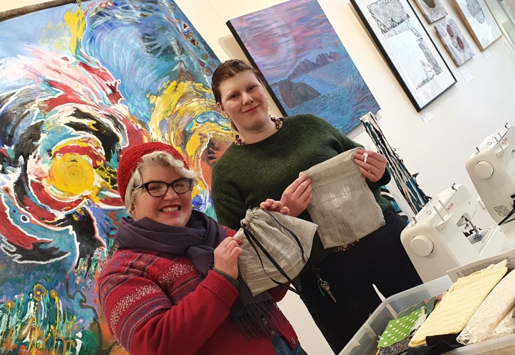 Two people smiling while holding handmade pouches. Theyre in a room with vibrant abstract art on the walls and sewing machines visible. One wears a red hat and glasses, the other a green sweater. A table with fabric is in the foreground.